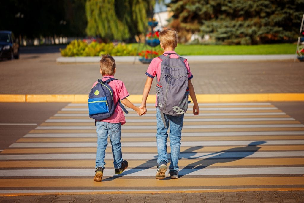 Two young boys wearing backpacks crossing street at crosswalk holding hands.