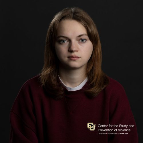 Headshot of a white person with shoulder-length red hair wearing a maroon sweatshirt and white tee.