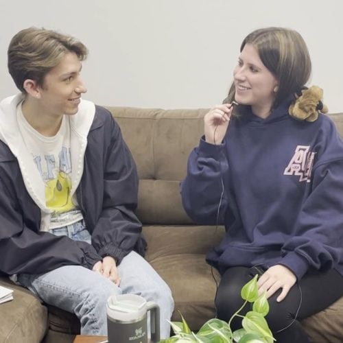Person with long brown hair in navy sweatshirt interviewing person with short brown hair in black windbreaker and white graphic tee.