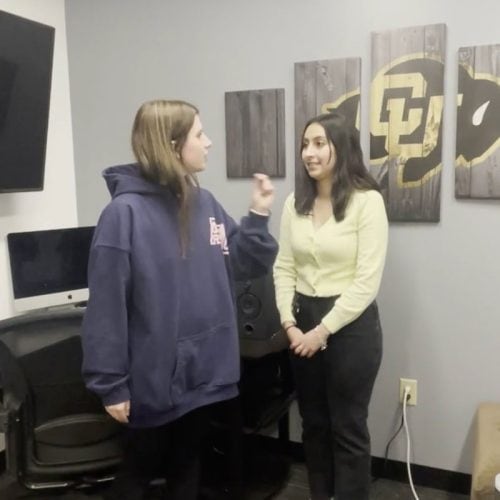 Person with long brown hair in navy sweatshirt interviewing person with long dark brown hair in pale yellow shirt.