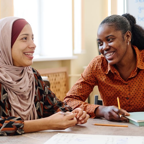 A Muslim woman and black woman sit at table with paper and pencils